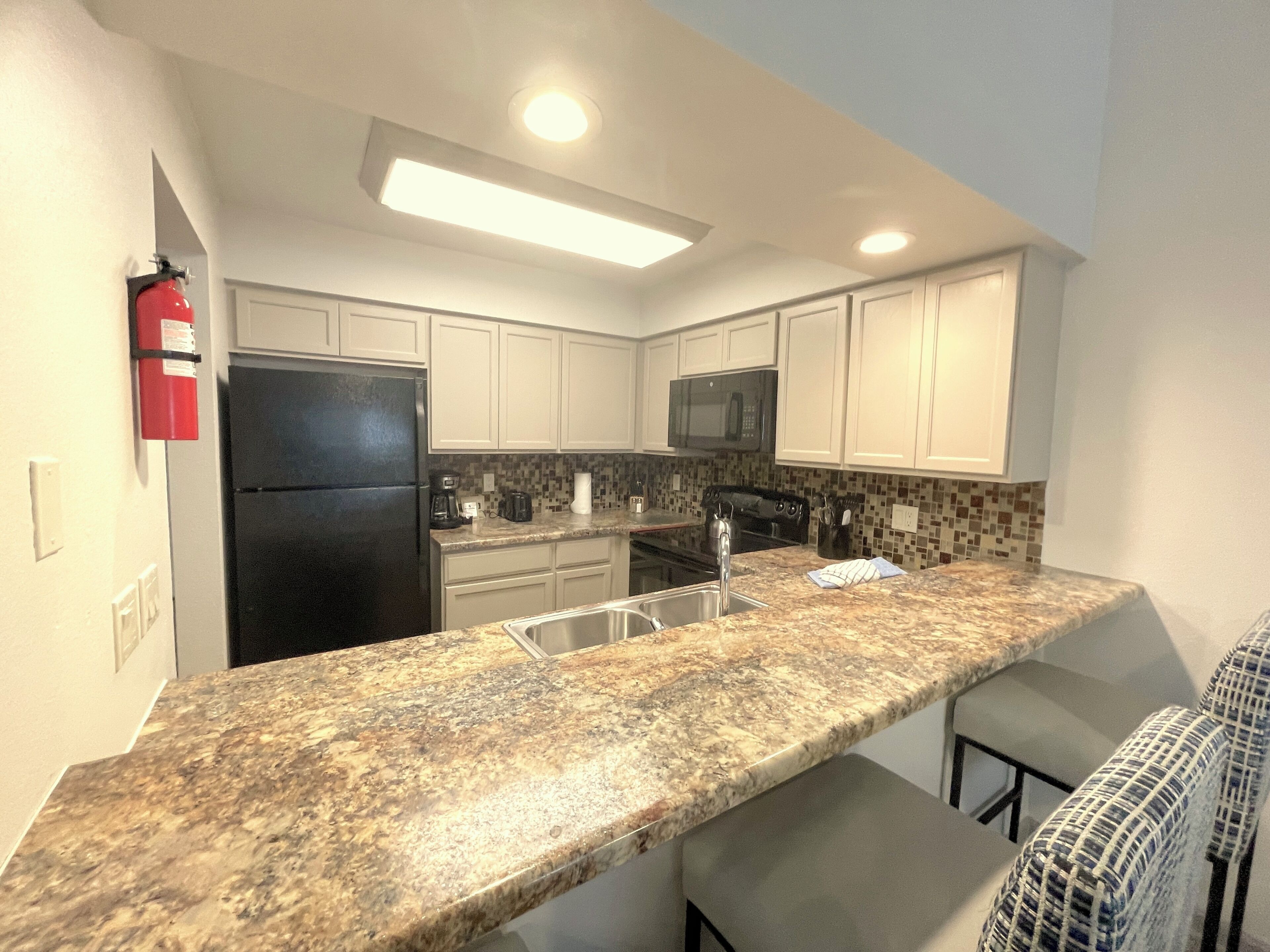 Una cocina con gabinetes blancos, un refrigerador negro, un microondas, una encimera de granito y un taburete de bar.