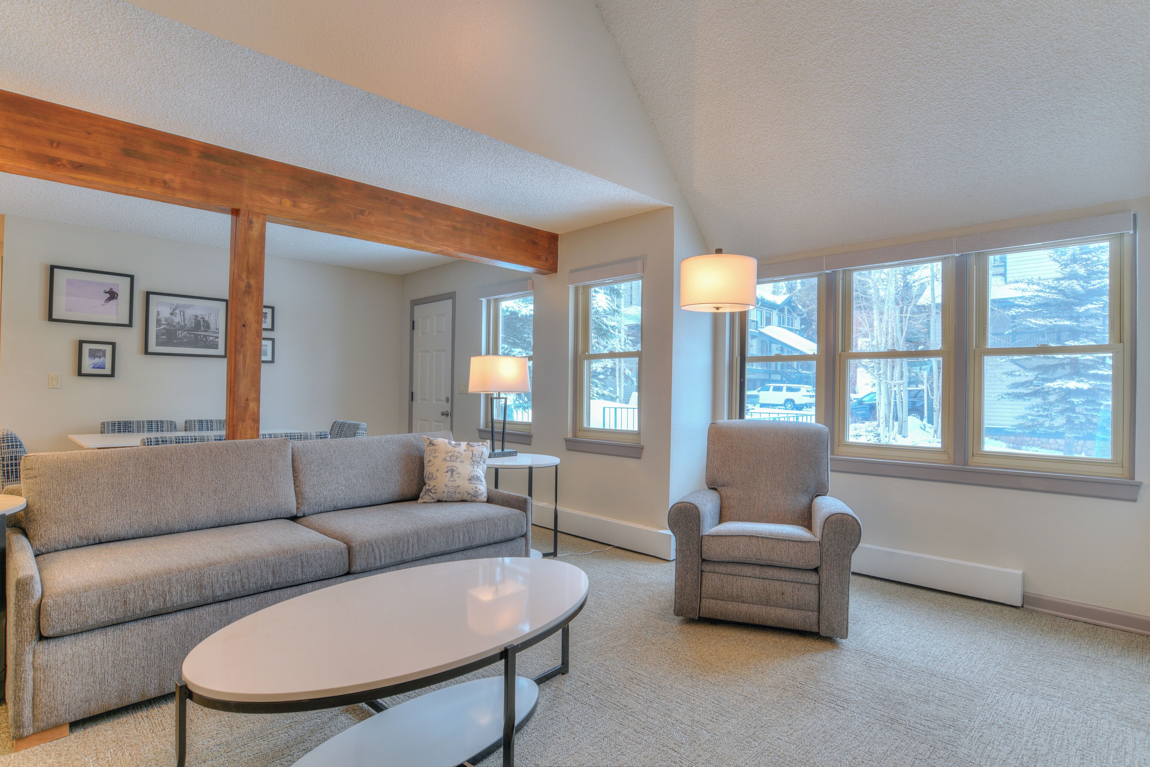 A living room with a sofa, armchair, and coffee table, large windows with a view of trees, and a wooden beam ceiling.
