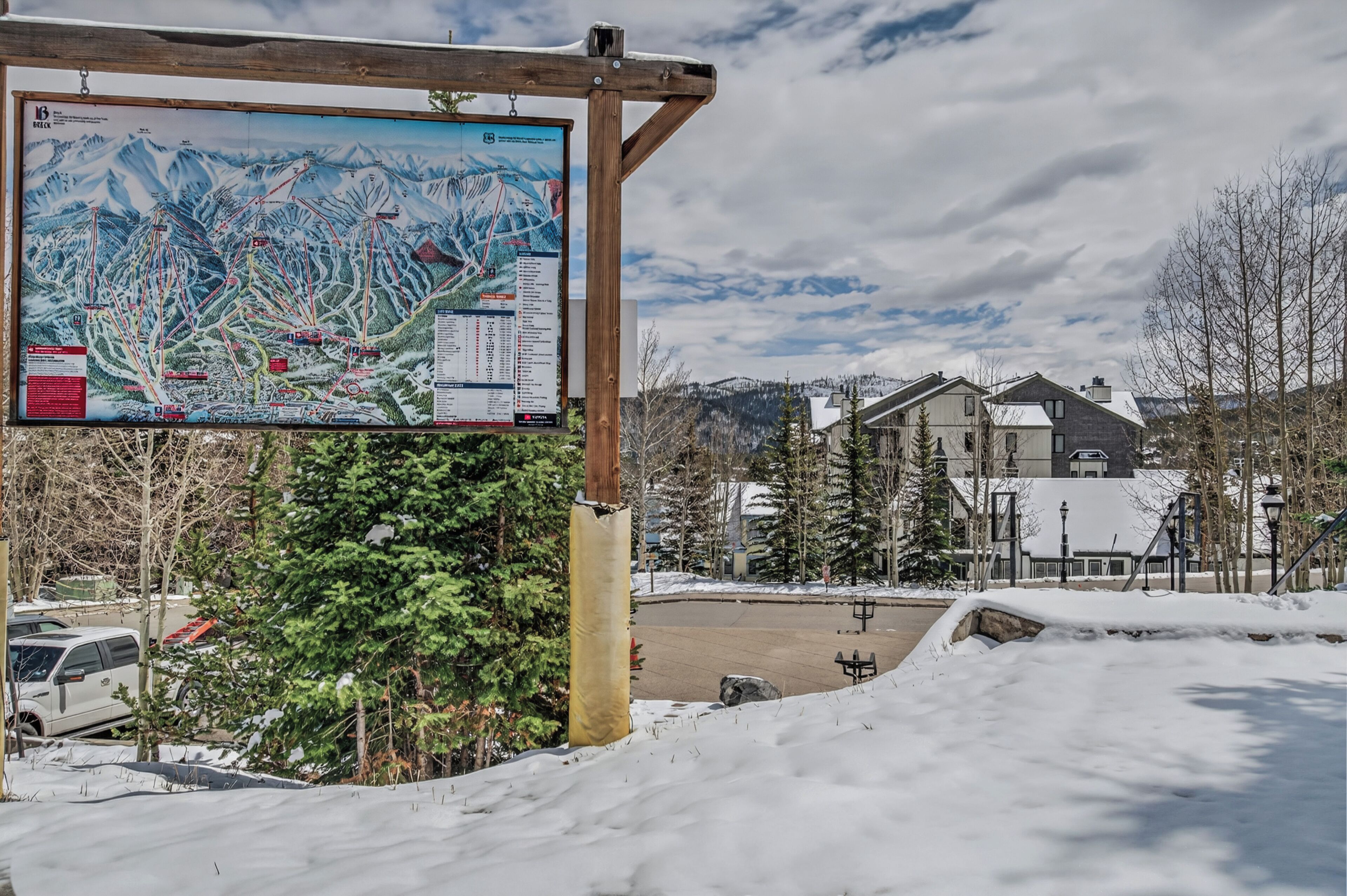 A ski resort with a large informational sign, snow-covered ground, and buildings in the background.