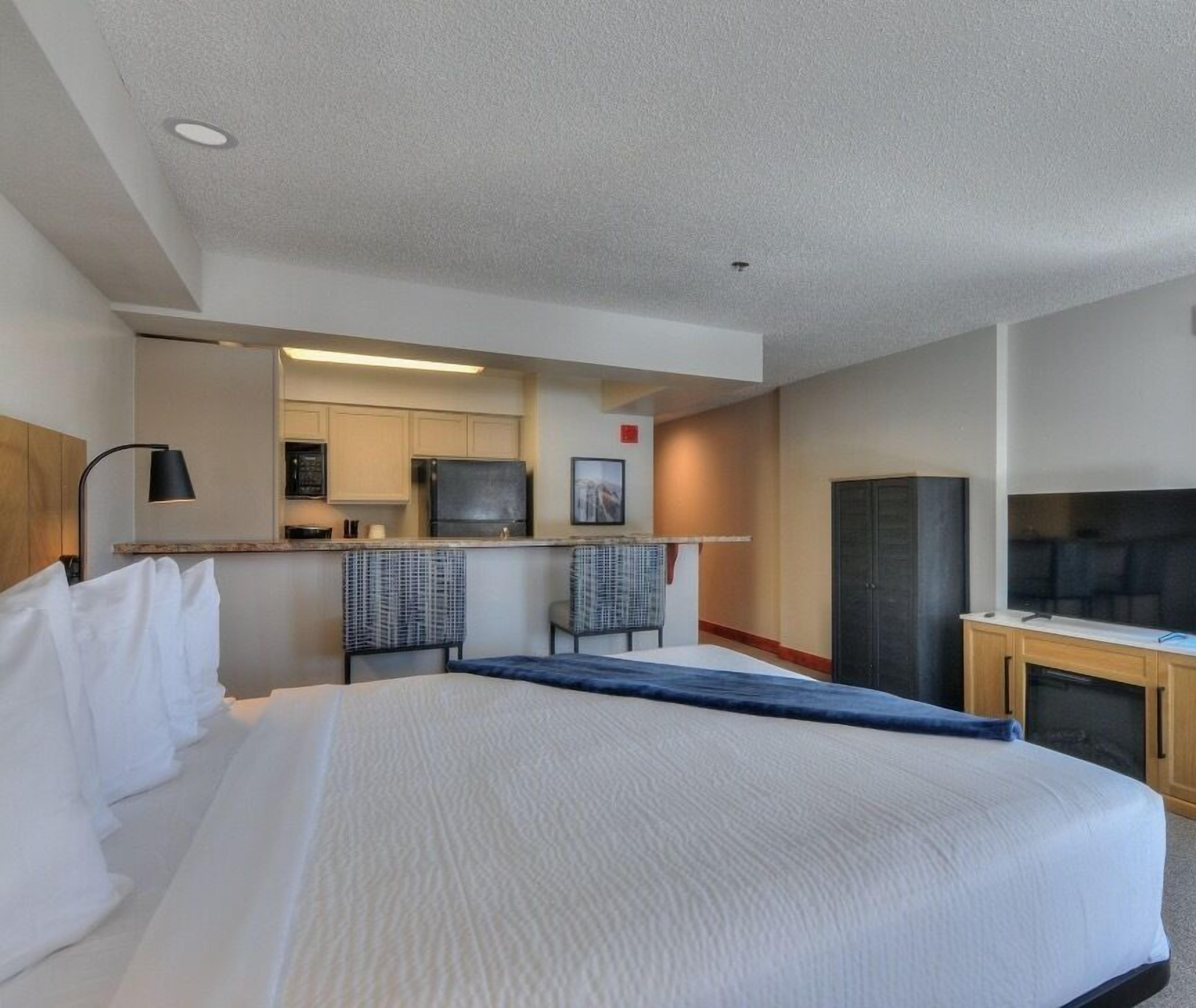 A hotel room with a large bed, a kitchenette, a TV, and a desk area.