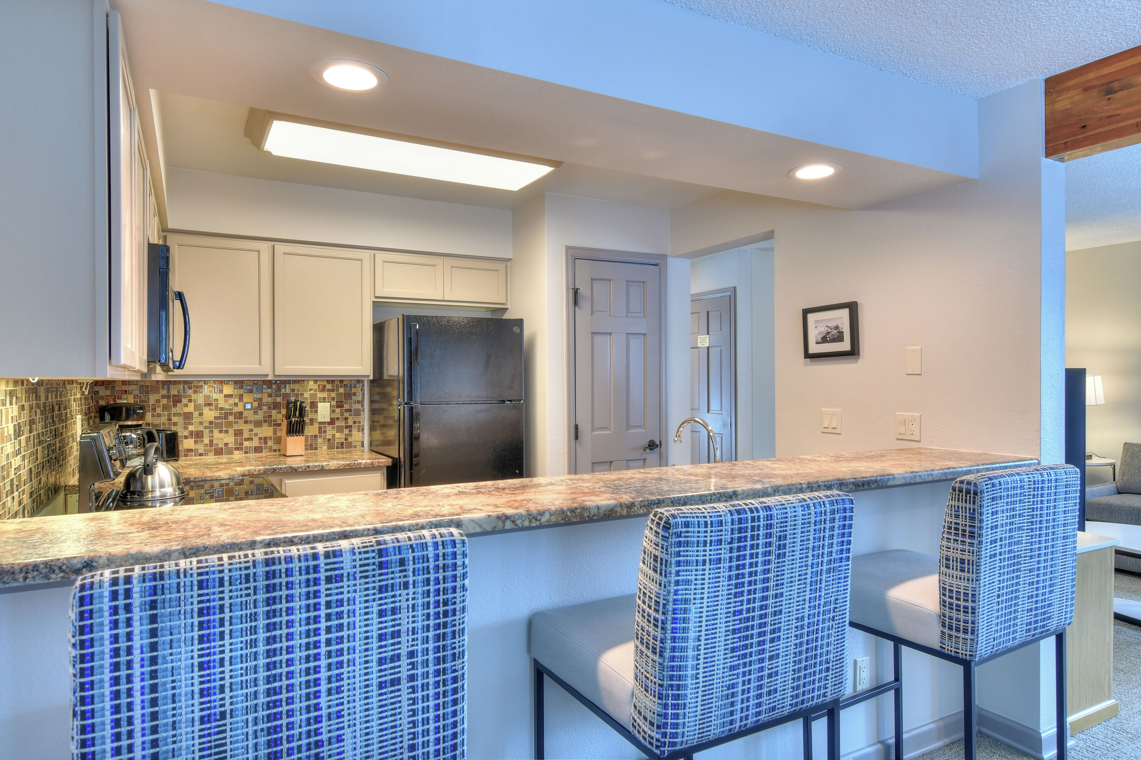 Cocina con armarios blancos, refrigerador de acero inoxidable, salpicadero de azulejos y una barra con sillas azules.