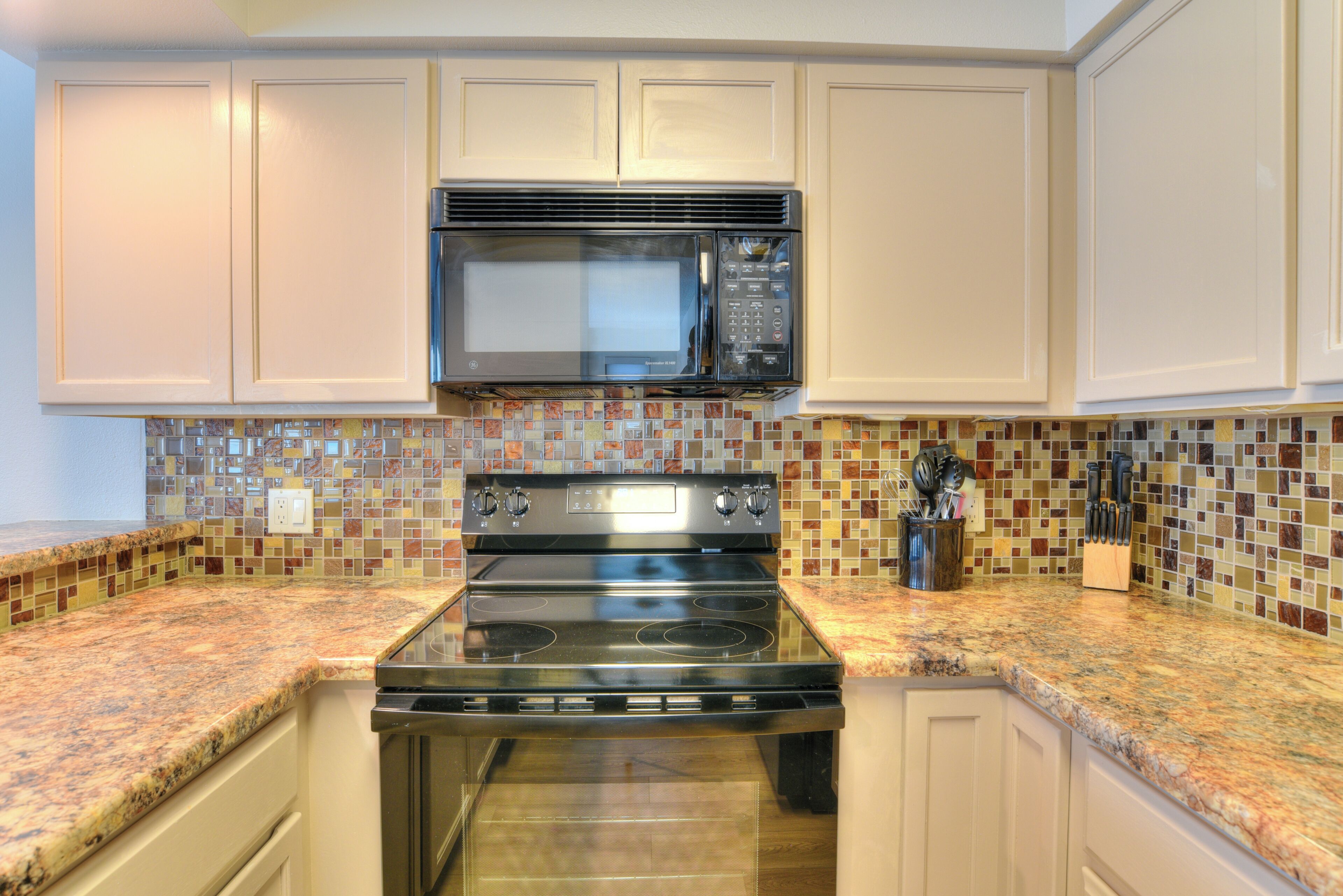 A kitchen with white cabinets, a black microwave, a stainless steel stove, and a colorful mosaic backsplash.
