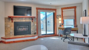 A living room with a fireplace, a sliding glass door leading outside, a desk with a chair, and a lamp. - Wedgewood Lodge (Breckenridge)