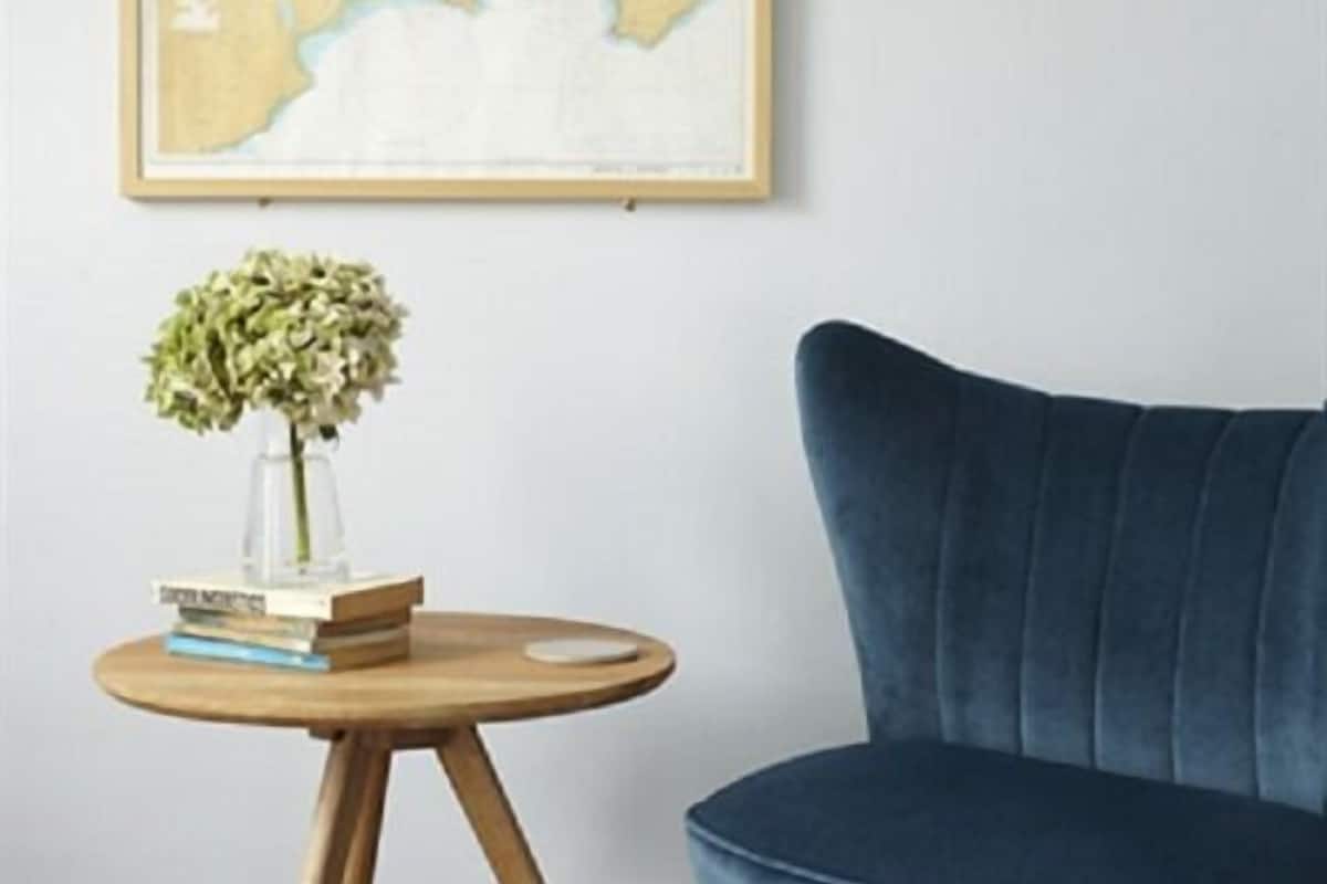 A room with a blue armchair, a round wooden side table with a vase of flowers and books, and a framed map on the wall.