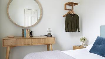 Un dormitorio con un escritorio de madera, un espejo redondo, una percha, una mesita de noche con lámpara y una cama con almohada azul.