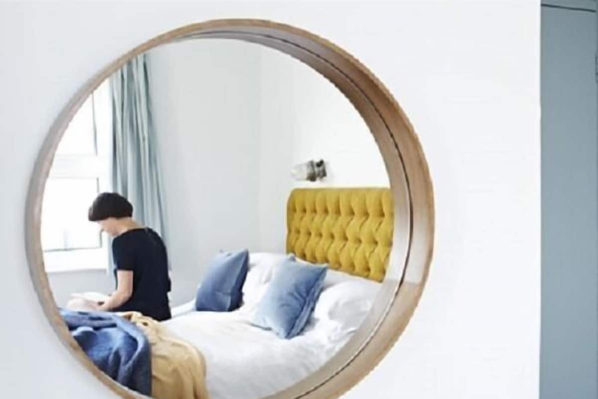 A round mirror reflecting a bed with a yellow headboard, white bedding, and blue pillows.