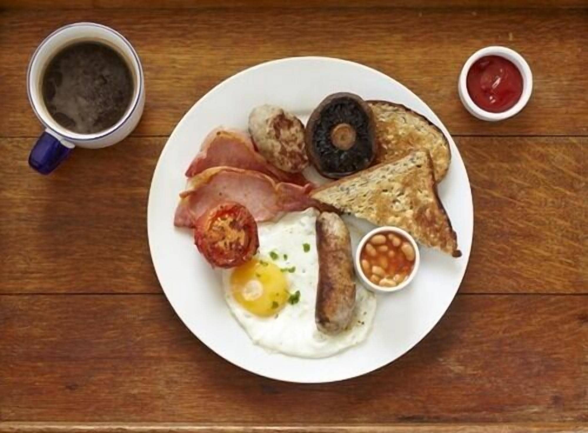 A plate of breakfast food including eggs, sausages, bacon, toast, and beans, with a cup of coffee and ketchup on the side.