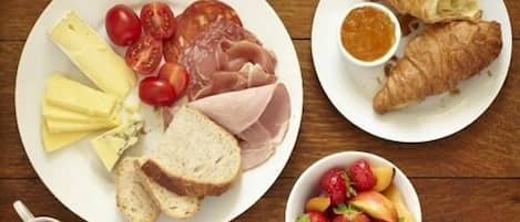 A breakfast plate with cheese, tomatoes, ham, bread, croissants, and a fruit bowl.