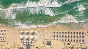 Une vue aérienne d’une plage avec des rangées de transats et de parasols, quelques personnes et un bâtiment au centre.