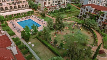 A large swimming pool surrounded by lounge chairs and umbrellas, with a playground nearby.