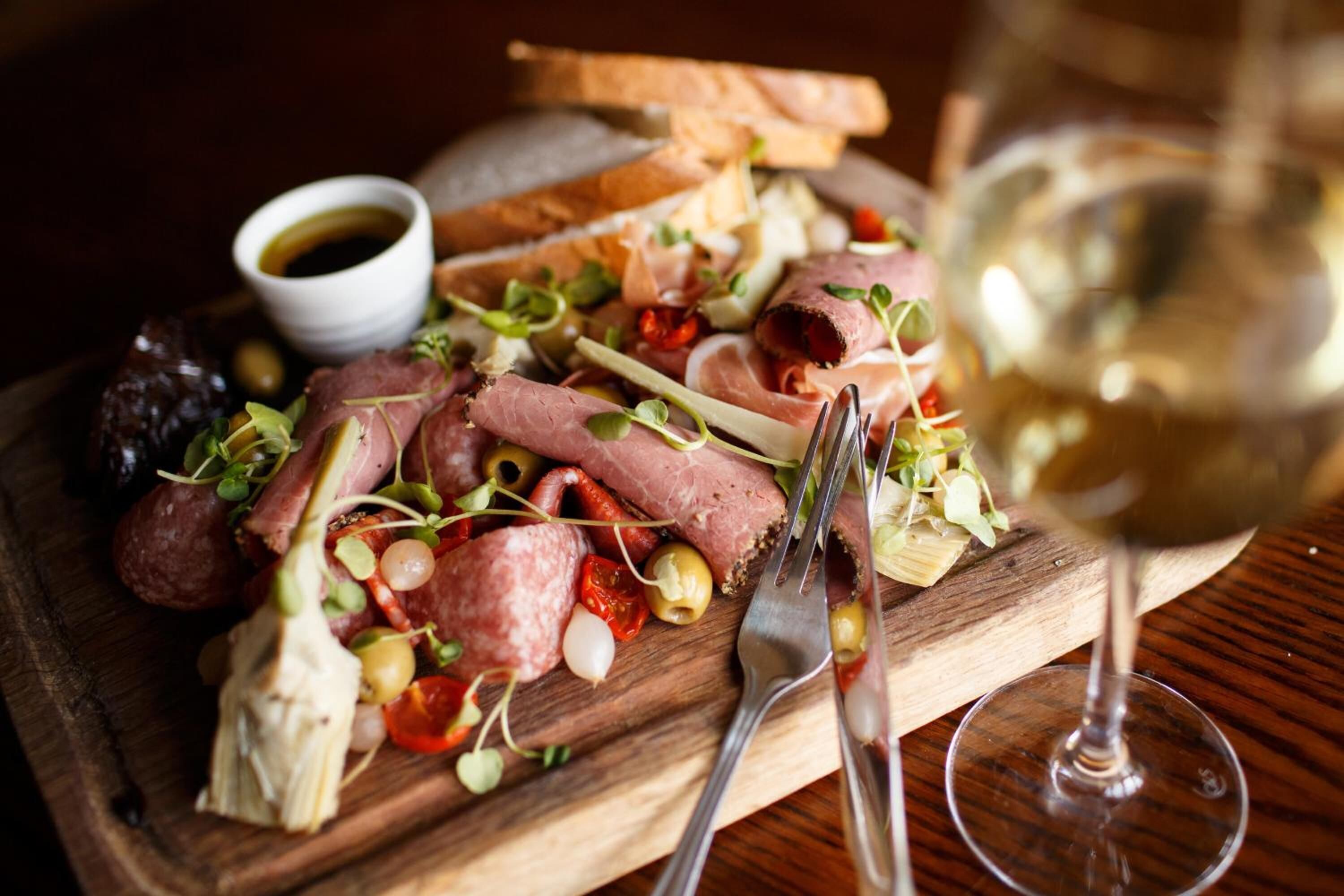 Une planche en bois garnie de différentes sortes de charcuterie, d’olives et de légumes verts, accompagnée d’un verre de vin blanc et d’un petit bol de sauce.