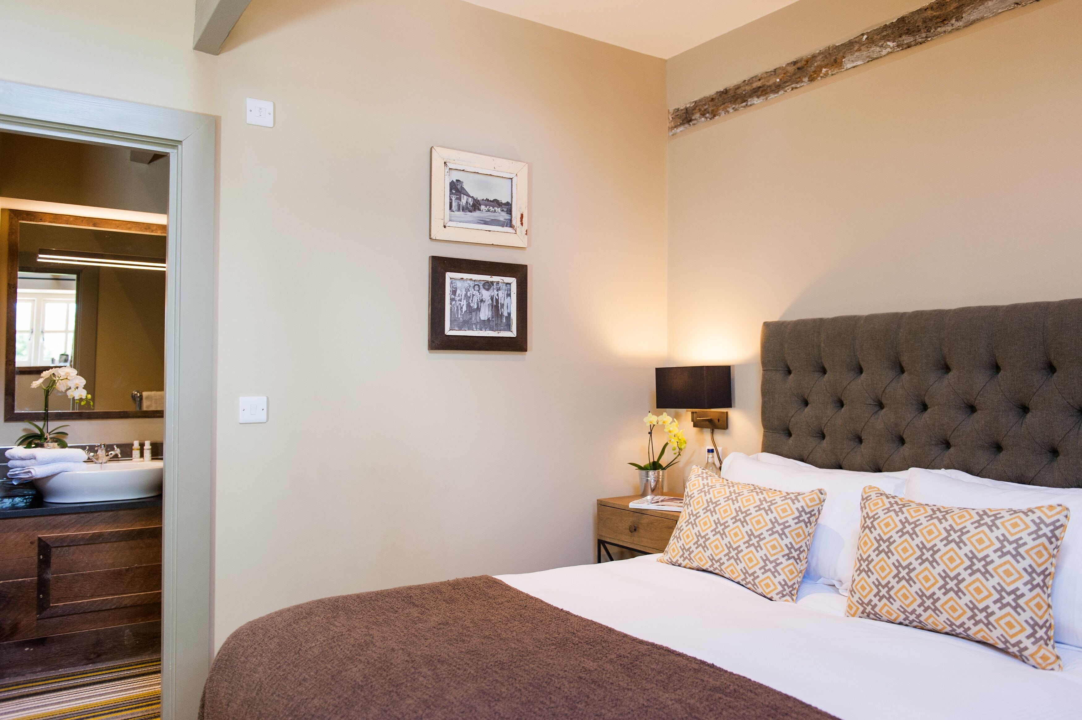 A hotel room with a large bed, a bedside table with a lamp, two framed pictures on the wall, and a bathroom visible through an open door.