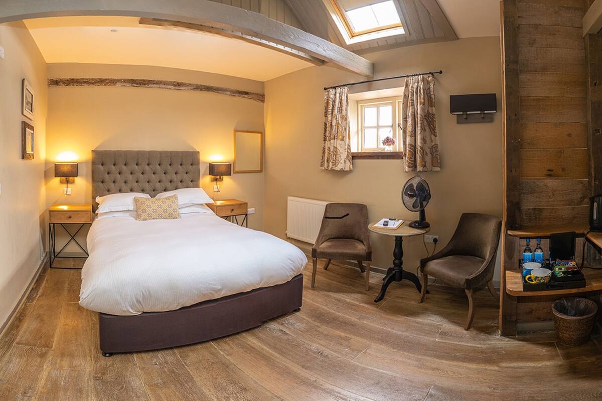 A bedroom with a large bed, two bedside tables with lamps, a chair, a small table with a fan, and a window with curtains.