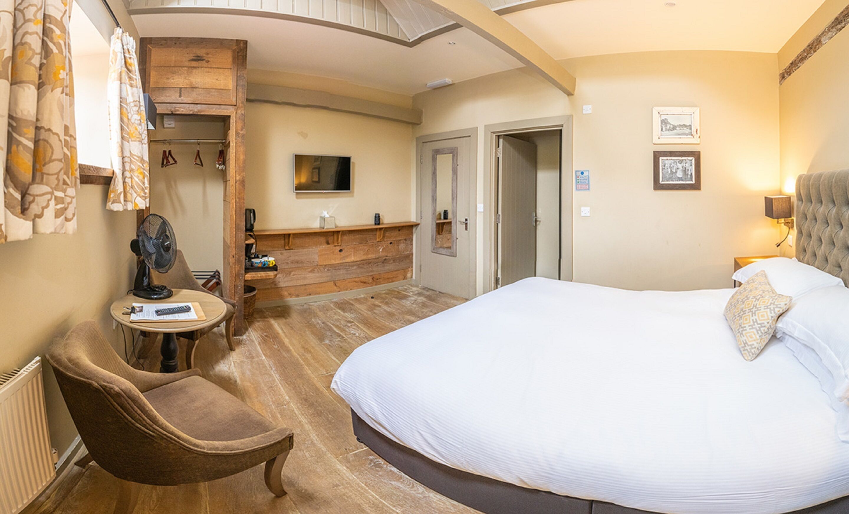 A hotel room with a large bed, a wooden vanity, a chair, a desk with a fan, and a television.