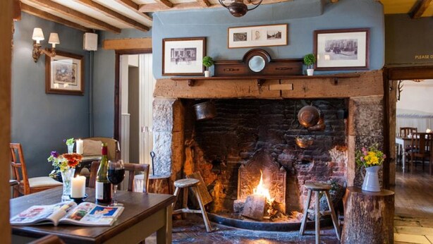 A cozy room with a stone fireplace, wooden beams, and framed pictures on the wall. - The White Hart, South Harting (Petersfield)