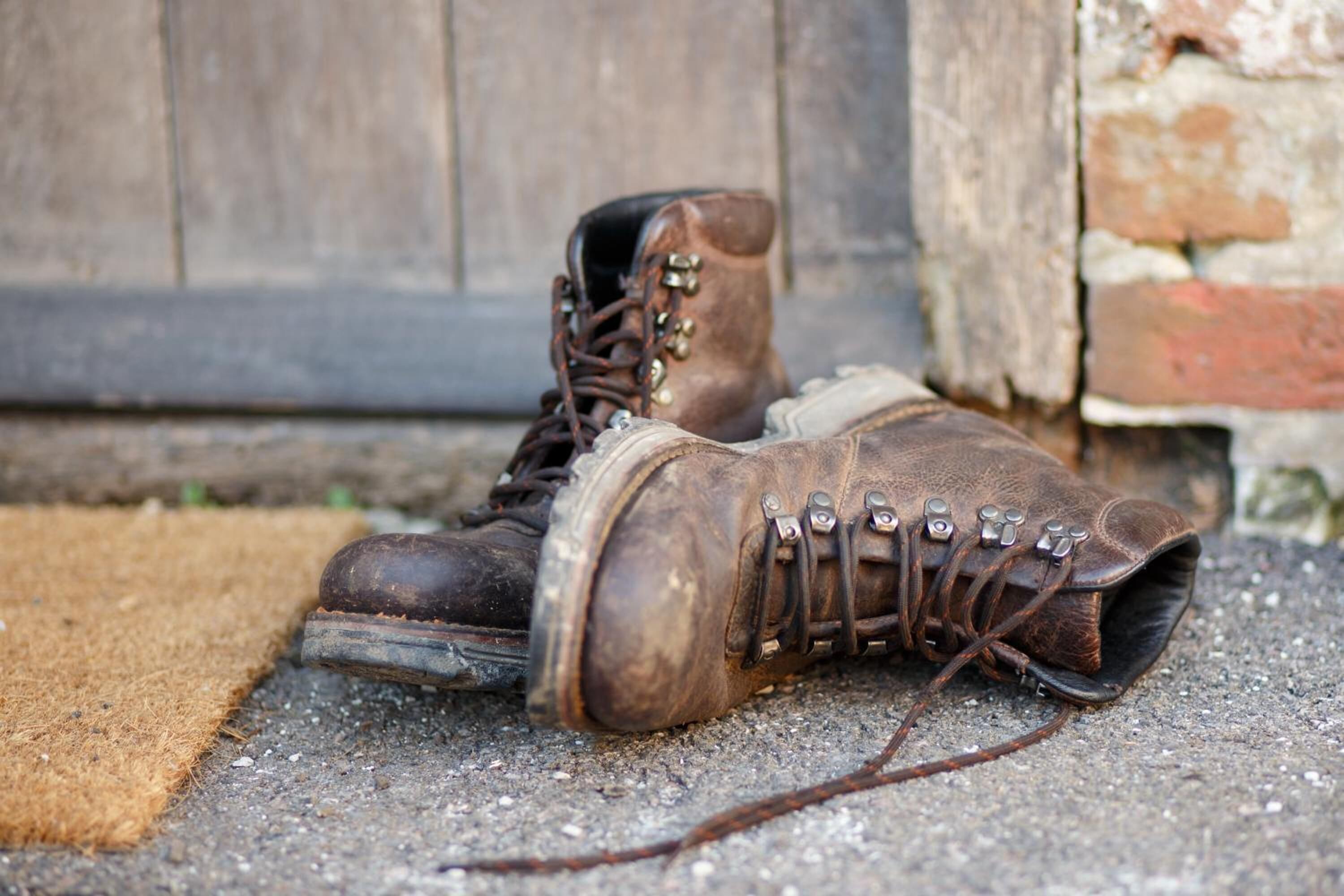Une paire de bottes marron usées, dotées d’œillets et de lacets en métal, est posée sur une surface en béton, à côté d’une porte en bois et d’un mur de briques.