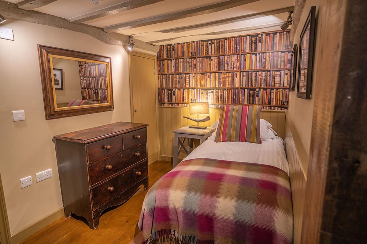 A cozy bedroom with a wooden dresser, a large bookshelf, and a bed with a plaid blanket.