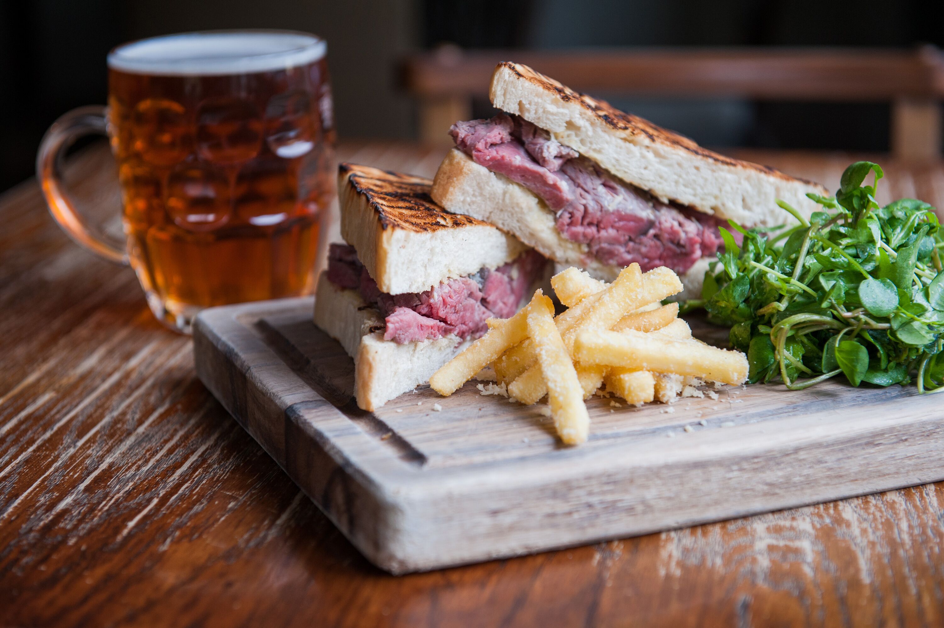 Un sandwich accompagné de frites et d’un verre de bière, le tout servi sur une planche en bois.