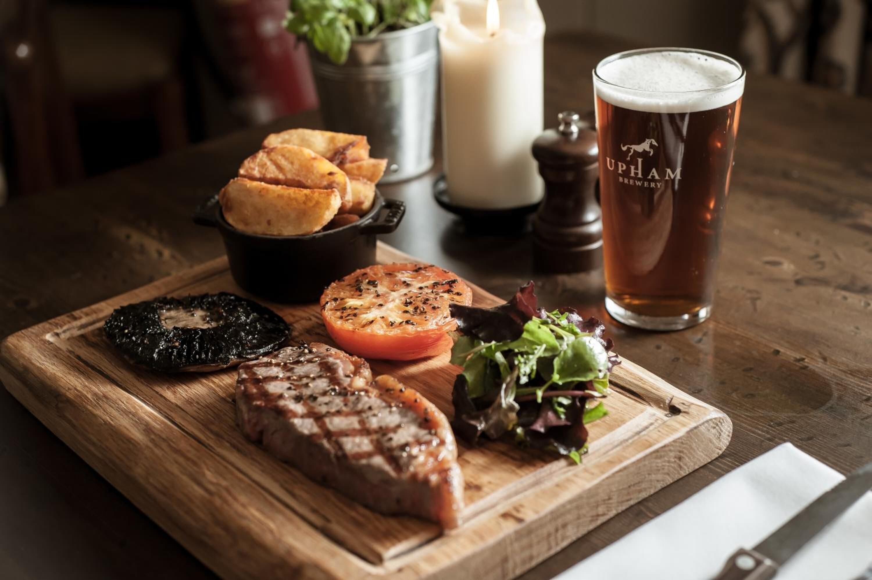 Una mesa de madera con un plato de carne a la parrilla, tomate asado, ensalada y plátanos fritos, acompañado de una cerveza.