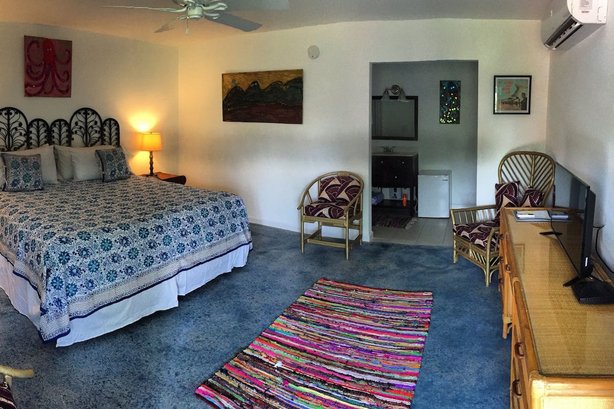 Quarto com cama, duas luminárias de cabeceira, ventilador de teto, tapete colorido e cômoda.