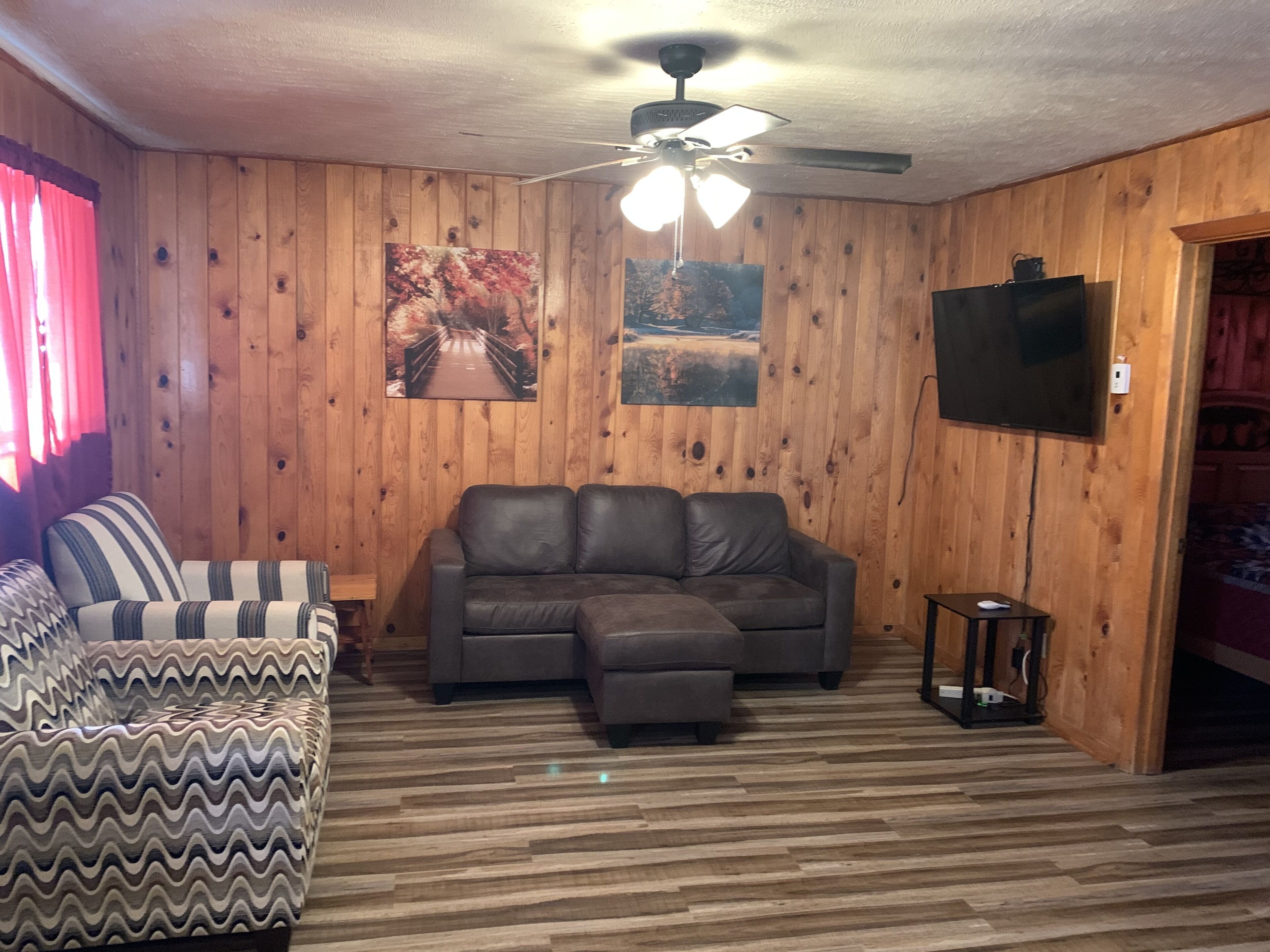 A room with wooden walls, a ceiling fan, a sectional sofa, a patterned armchair, and a television.