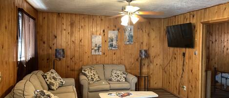 A cozy living room with wooden walls, a ceiling fan, a flat-screen TV, two sofas with patterned cushions, a coffee table, and a patterned rug.