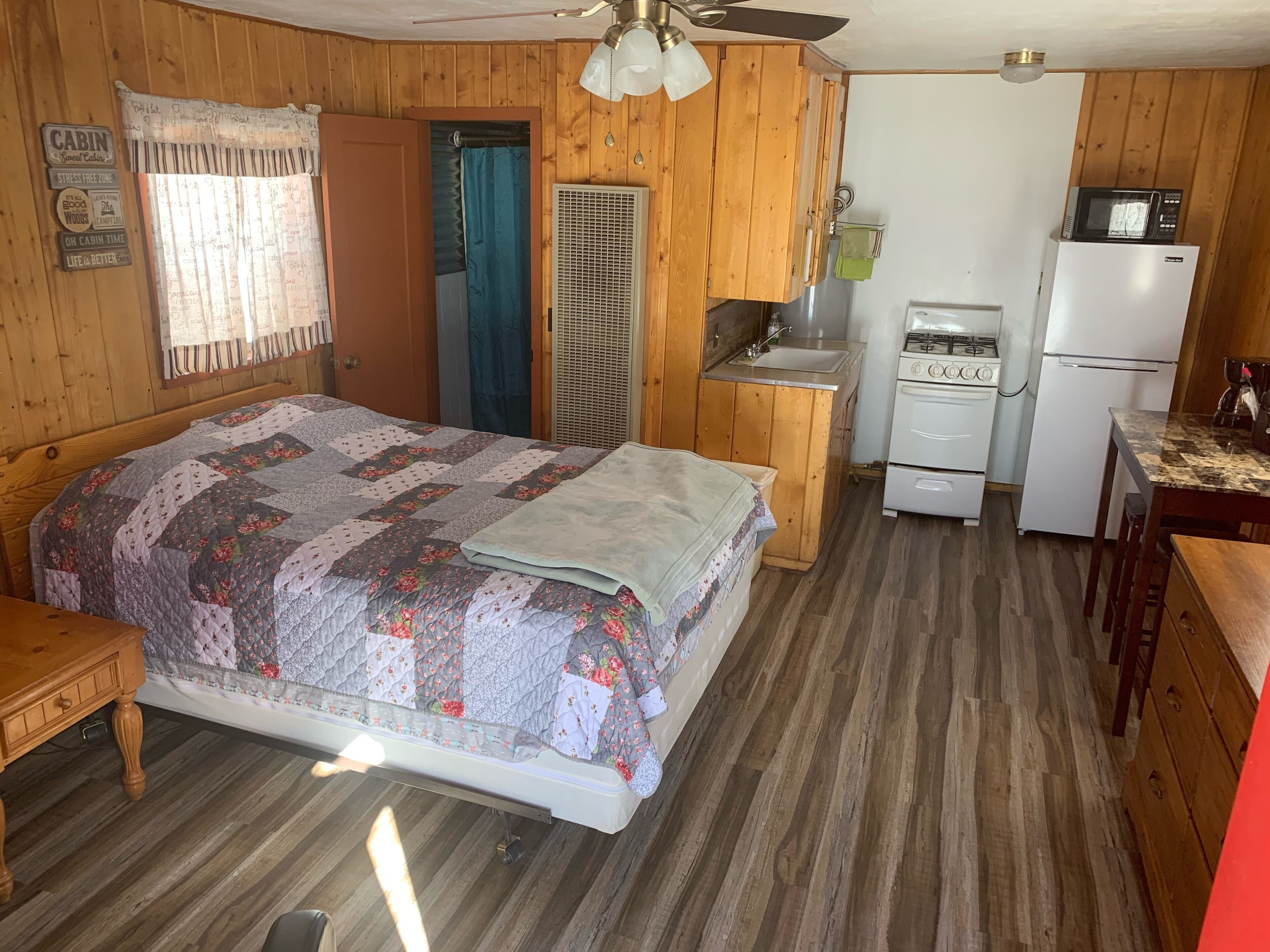 Una habitación tipo cabaña con cama, kitchenette, ventilador de techo y ventana con cortinas.