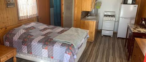 Una habitación tipo cabaña con cama, kitchenette, ventilador de techo y ventana con cortinas.