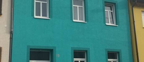 A multi-story building with a teal facade, white-framed windows, and a white door.