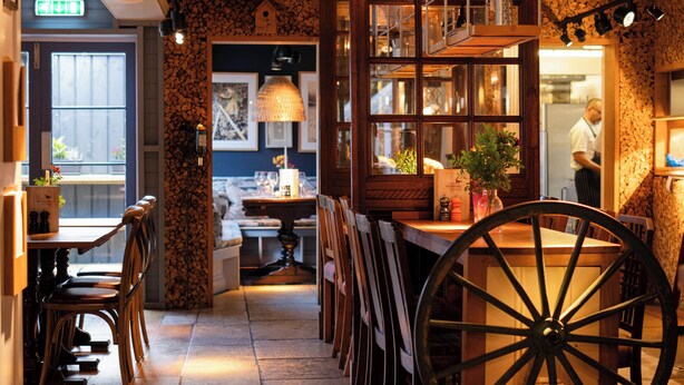 A narrow hallway with a large wooden wheel, tables, chairs, and a kitchen area visible in the background. - The Running Horse (Winchester)
