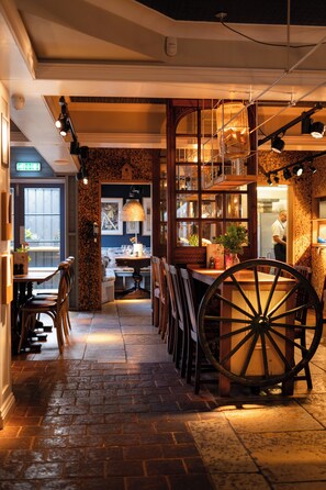 A narrow hallway with a large wooden wheel, tables, chairs, and a kitchen area visible in the background. - The Running Horse (Winchester)