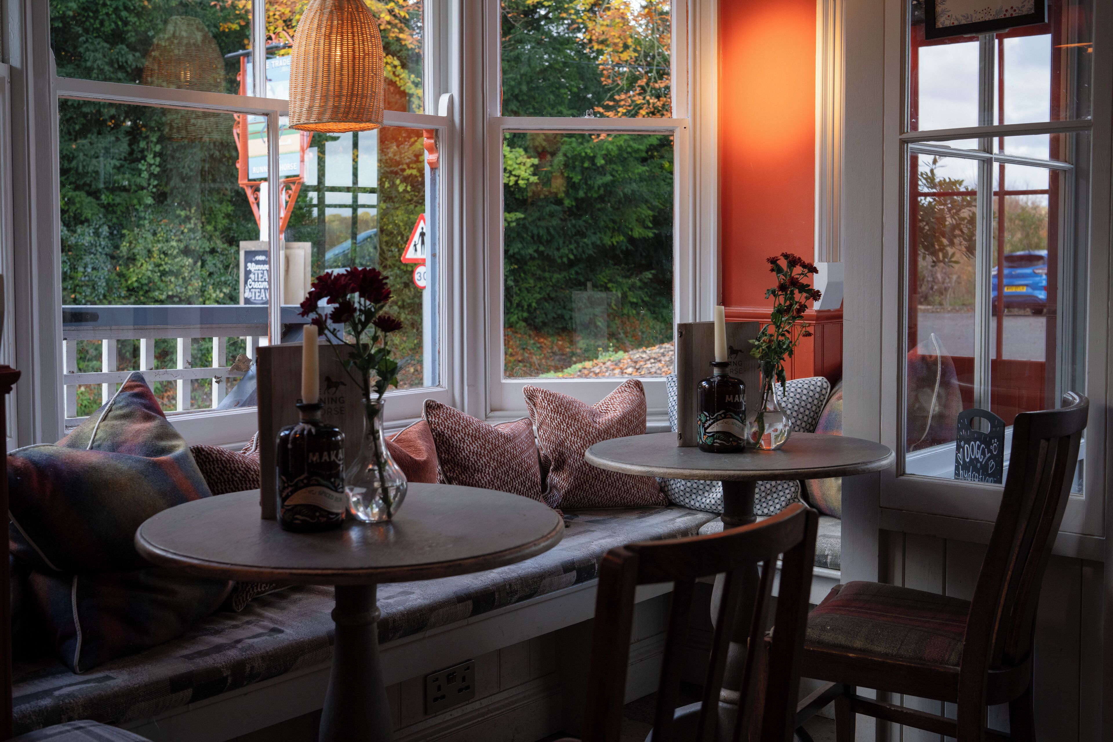 A cozy seating area with a bench and chairs, a round table with a bottle and vase, and large windows with a view of trees and a car outside.