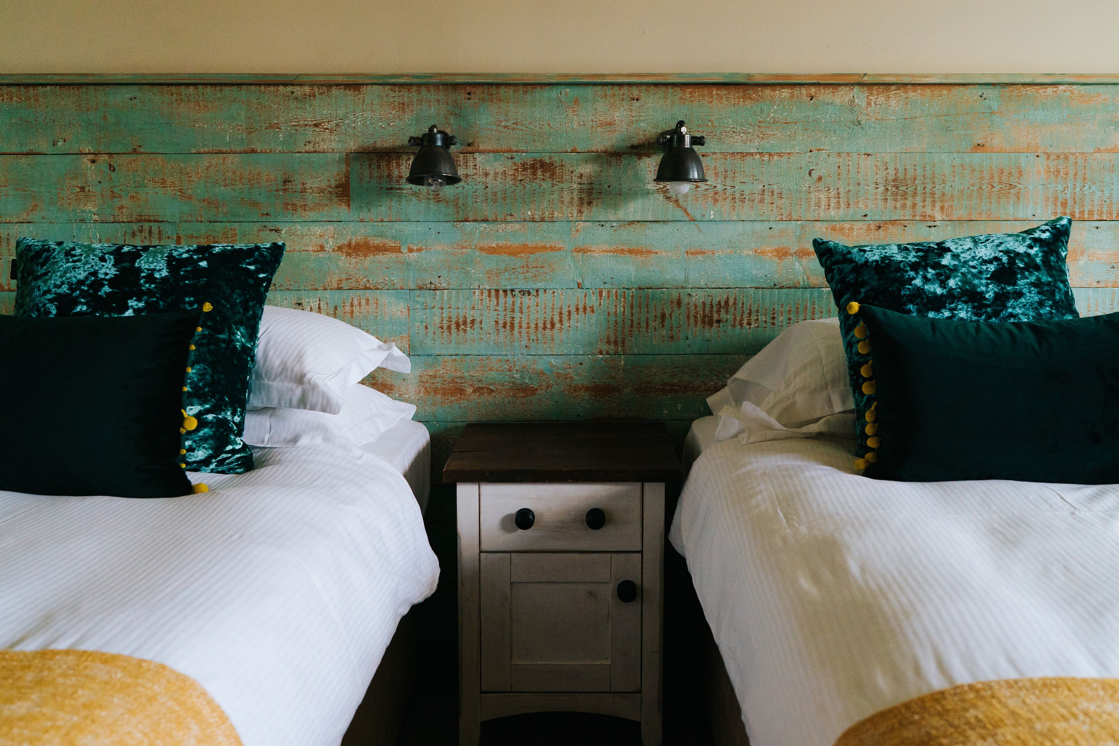 A room with two beds, each with white linens and dark green pillows. There is a wooden nightstand with two drawers and two wall-mounted lamps between the beds.