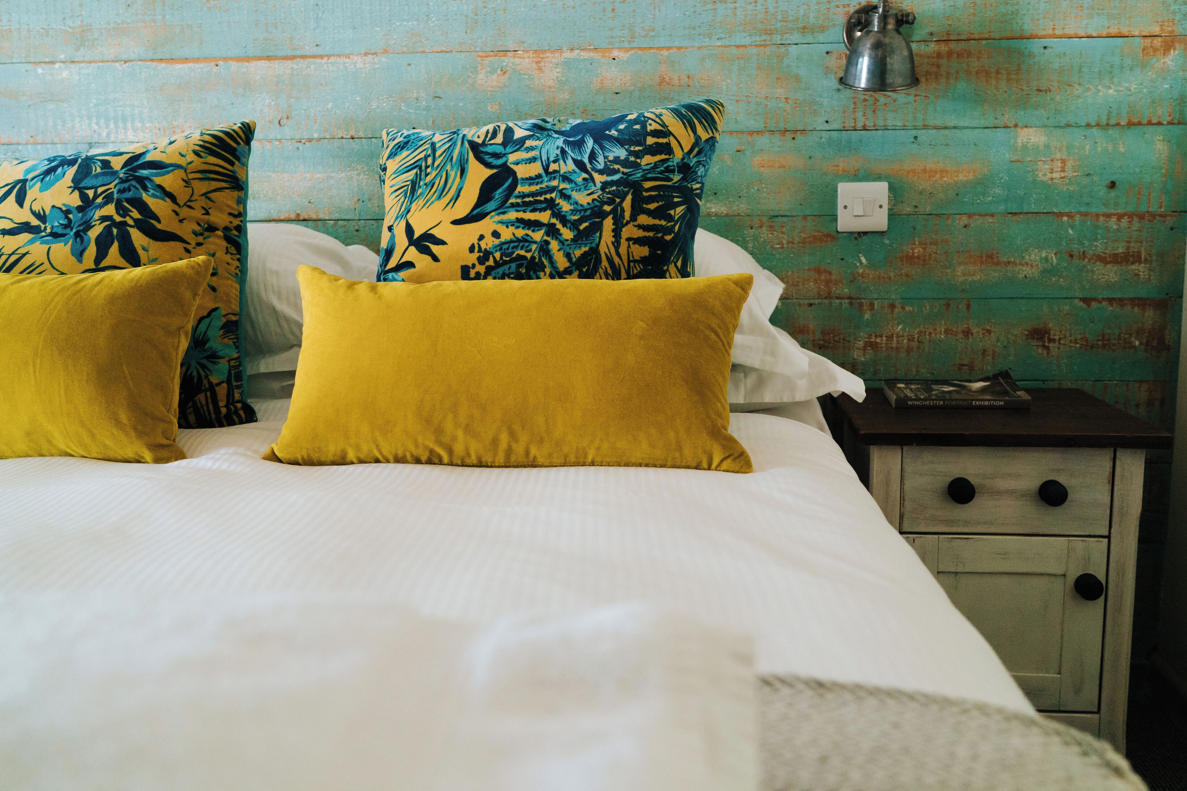 Un lit avec une literie blanche et deux oreillers jaunes, une table de chevet avec une lampe, et un mur peint en vert turquoise.