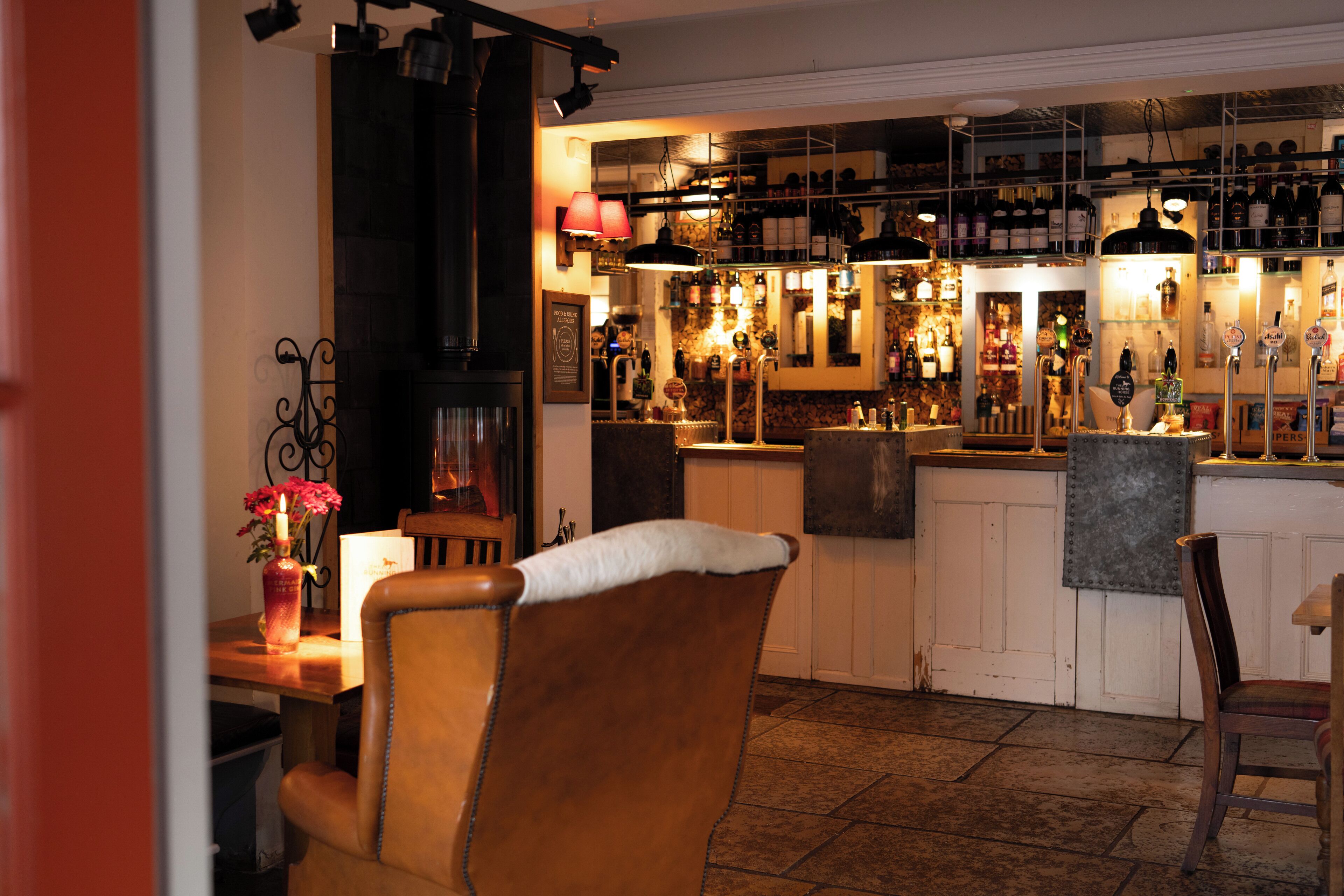 A bar area with a wooden counter, shelves of bottles, and a seating area with chairs.