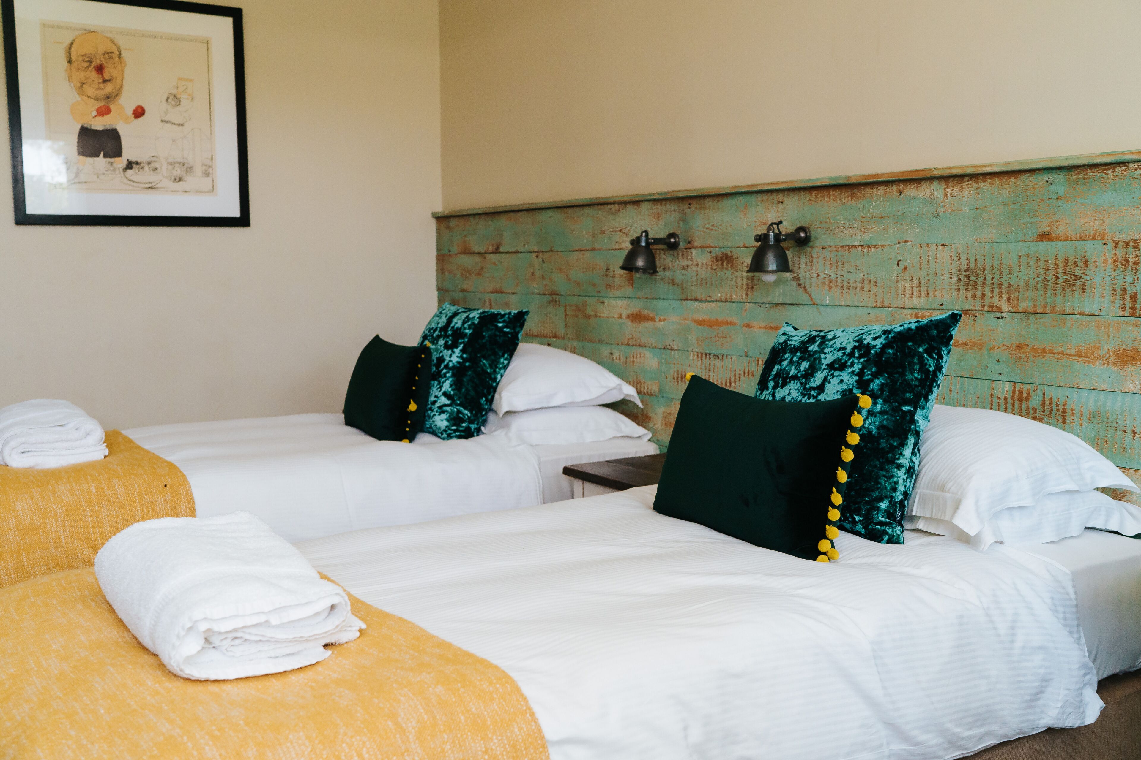 Une chambre d’hôtel avec deux lits, chacun étant équipé de draps blancs et d’oreillers verts. On trouve des serviettes jaunes sur les lits, et un tableau encadré est accroché au mur.