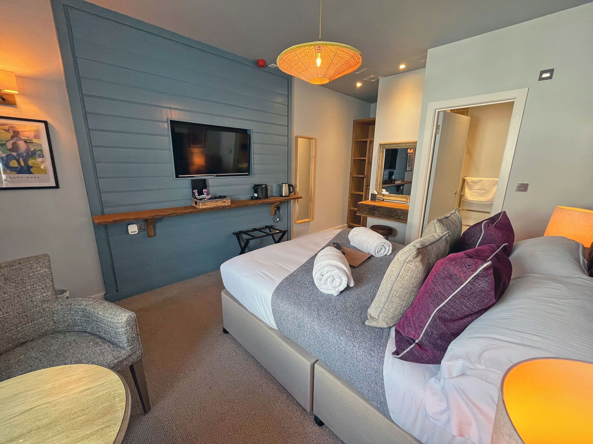 Habitación de hotel con cama, televisor, estante con artículos de aseo, una silla y un espejo.