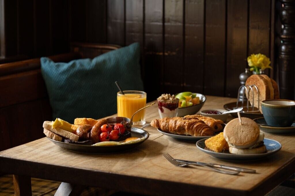 Un desayuno con cruasanes, tostadas, fruta y un vaso de zumo de naranja sobre una mesa de madera.