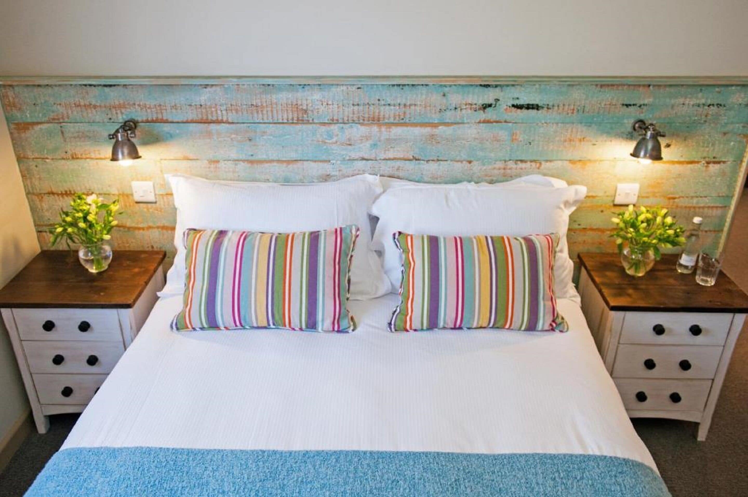 A bed with white linens, two striped pillows, and a blue throw. There are two nightstands with lamps and small vases with flowers.