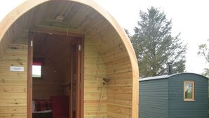 A wooden cabin with a curved entrance, a small porch, and a separate green building in the background. - Siabod Huts (Llanrwst)