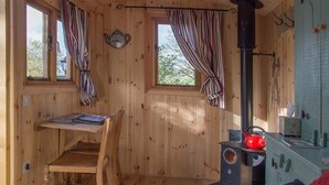 A cozy cabin interior with wooden walls, a desk, a chair, a fur rug, a stove, and a window with curtains. - Siabod Huts (Llanrwst)