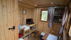 A wooden cabin interior with a kitchenette, a dining area, and a window with curtains. - Siabod Huts (Llanrwst)