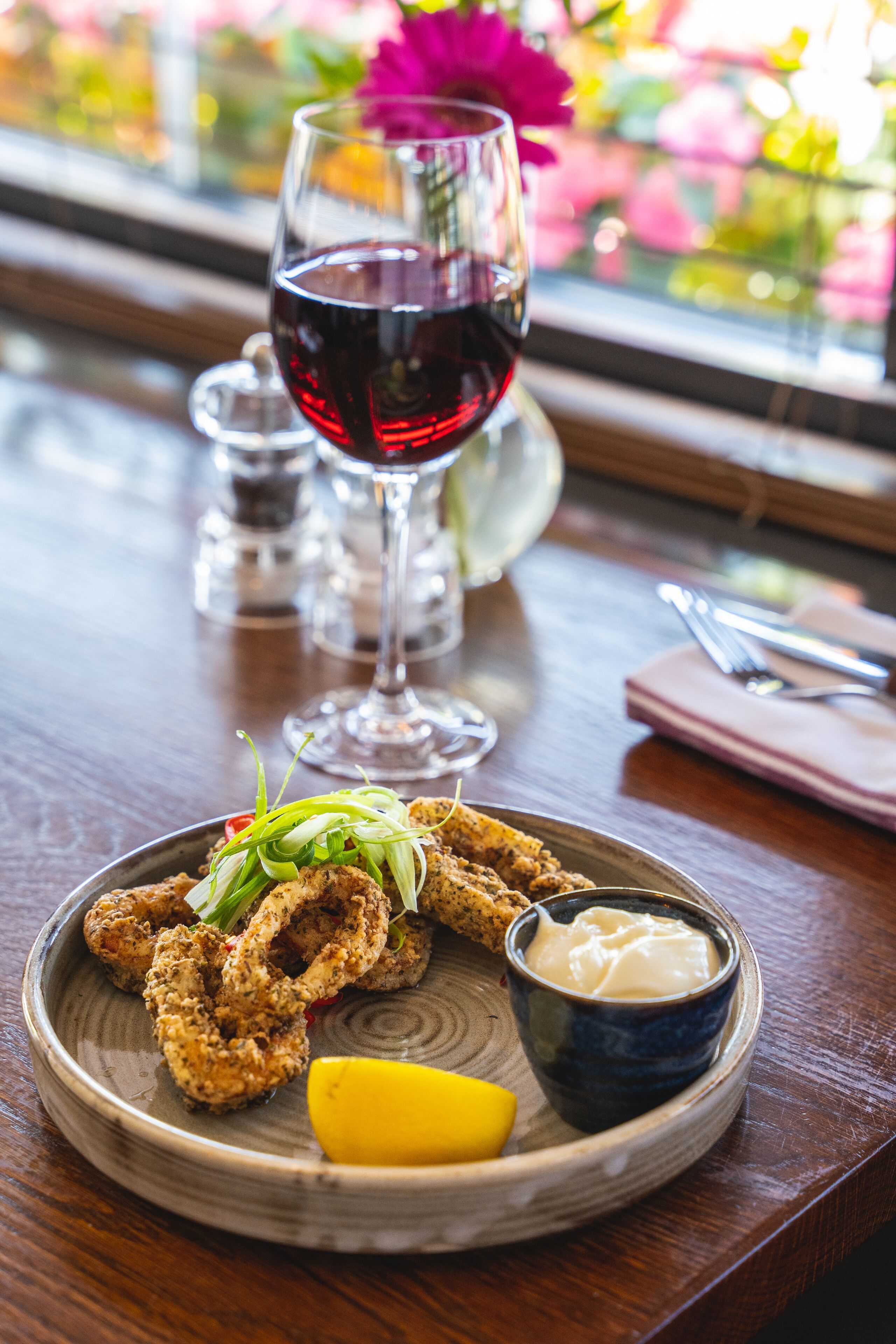 A plate of fried seafood with a lemon wedge and dipping sauce, a glass of red wine, and a window with flowers in the background.