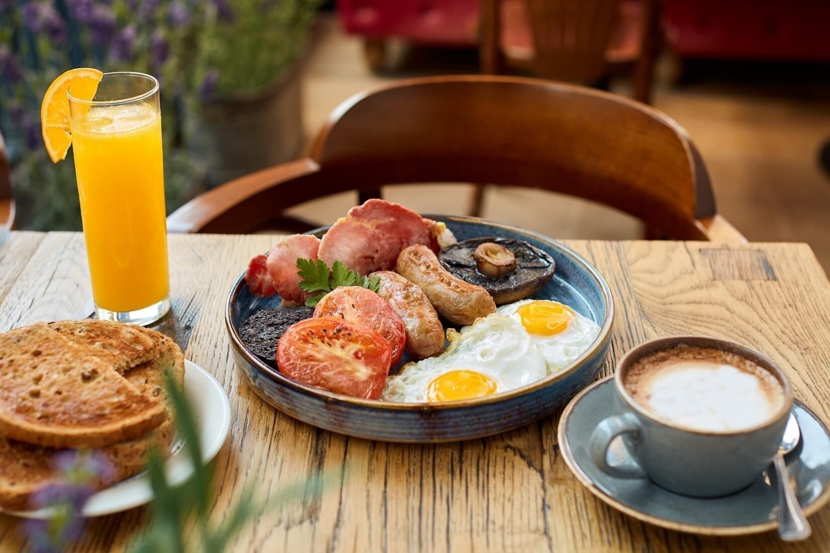 A breakfast plate with eggs, sausages, mushrooms, and toast, a glass of orange juice, and a cup of coffee on a wooden table.