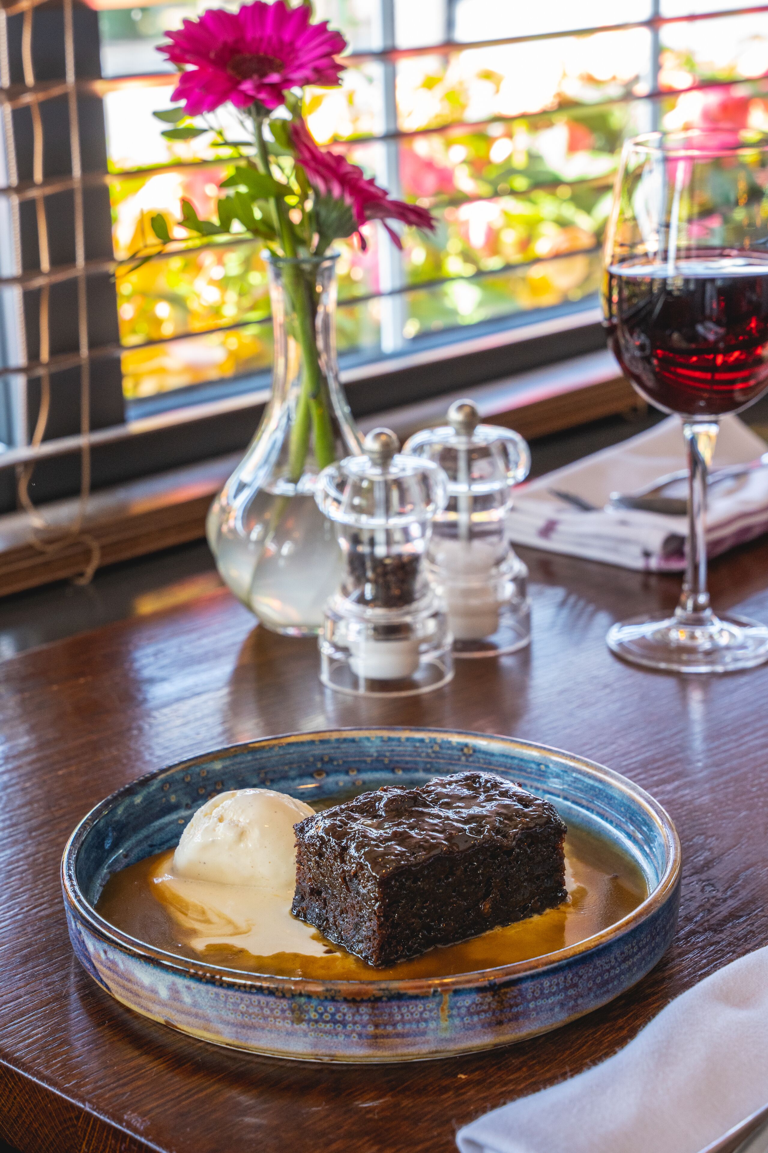 Un plato de postre con un pastel de chocolate y helado, una copa de vino tinto y un jarrón con flores sobre una mesa de madera.