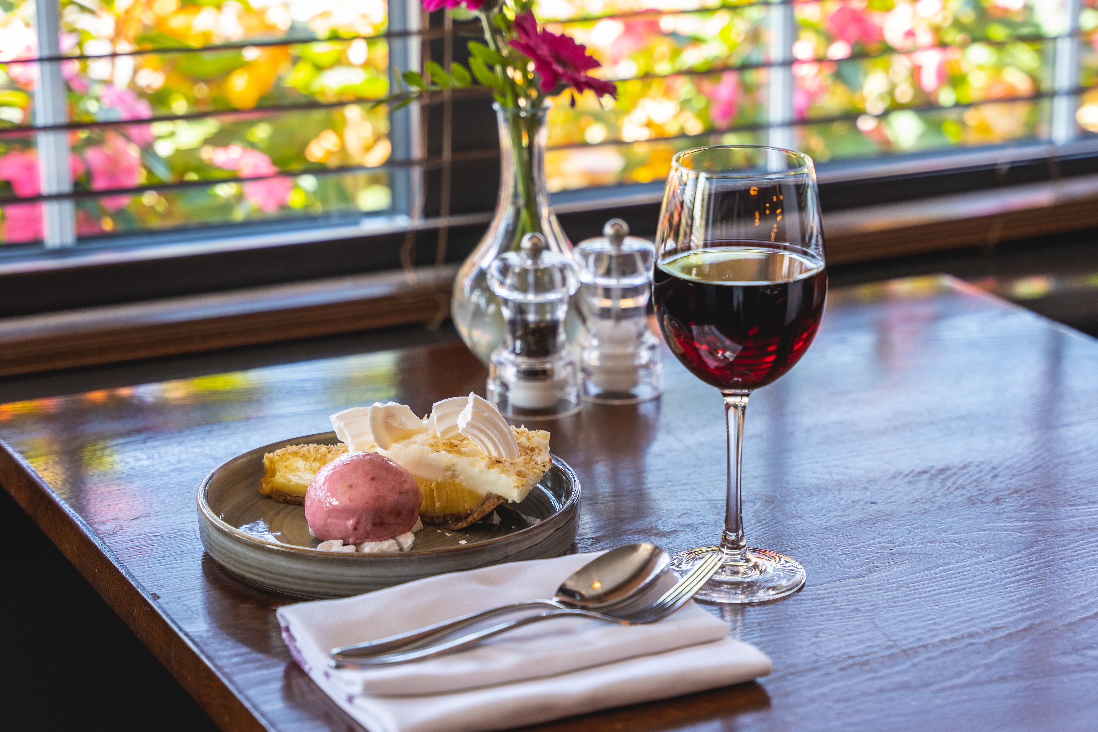 A dining table with a plate of dessert, a glass of red wine, and a vase with flowers.
