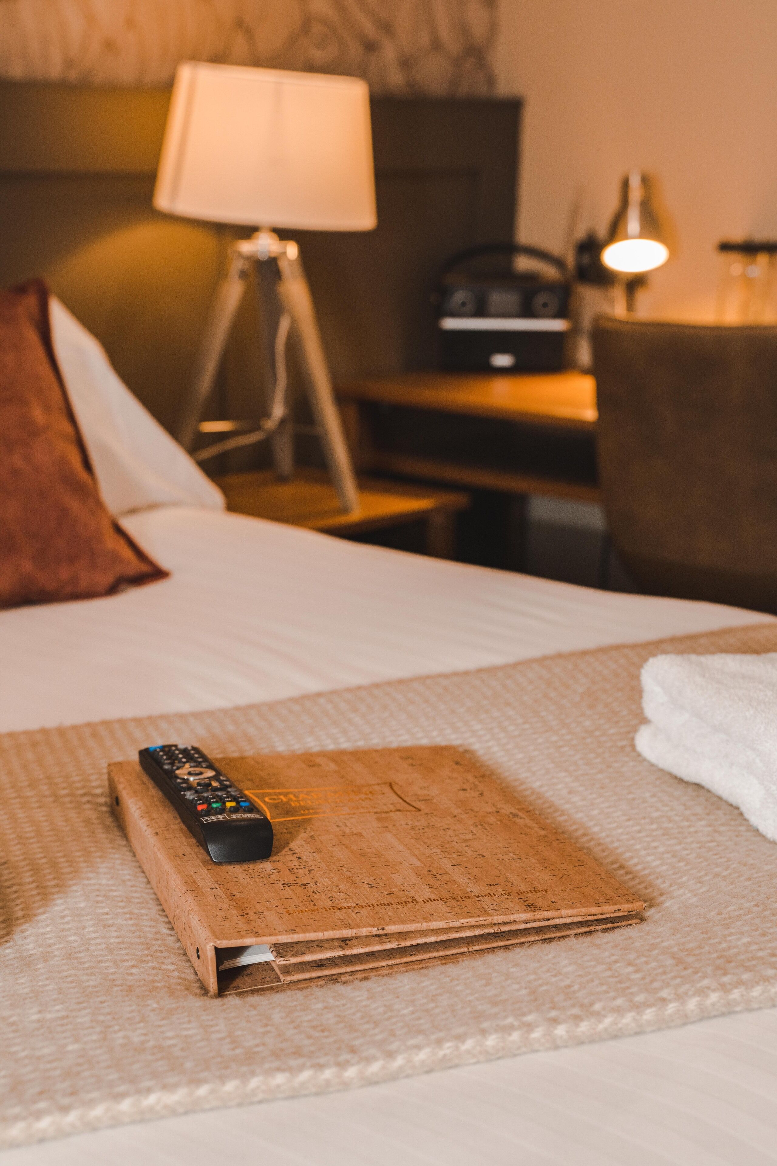 A hotel room with a bed, a remote control on a cork mat, a lamp, and a desk with a radio.