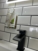 A bathroom with white tiles, a black faucet, and two bottles of hand sanitizer on a shelf.