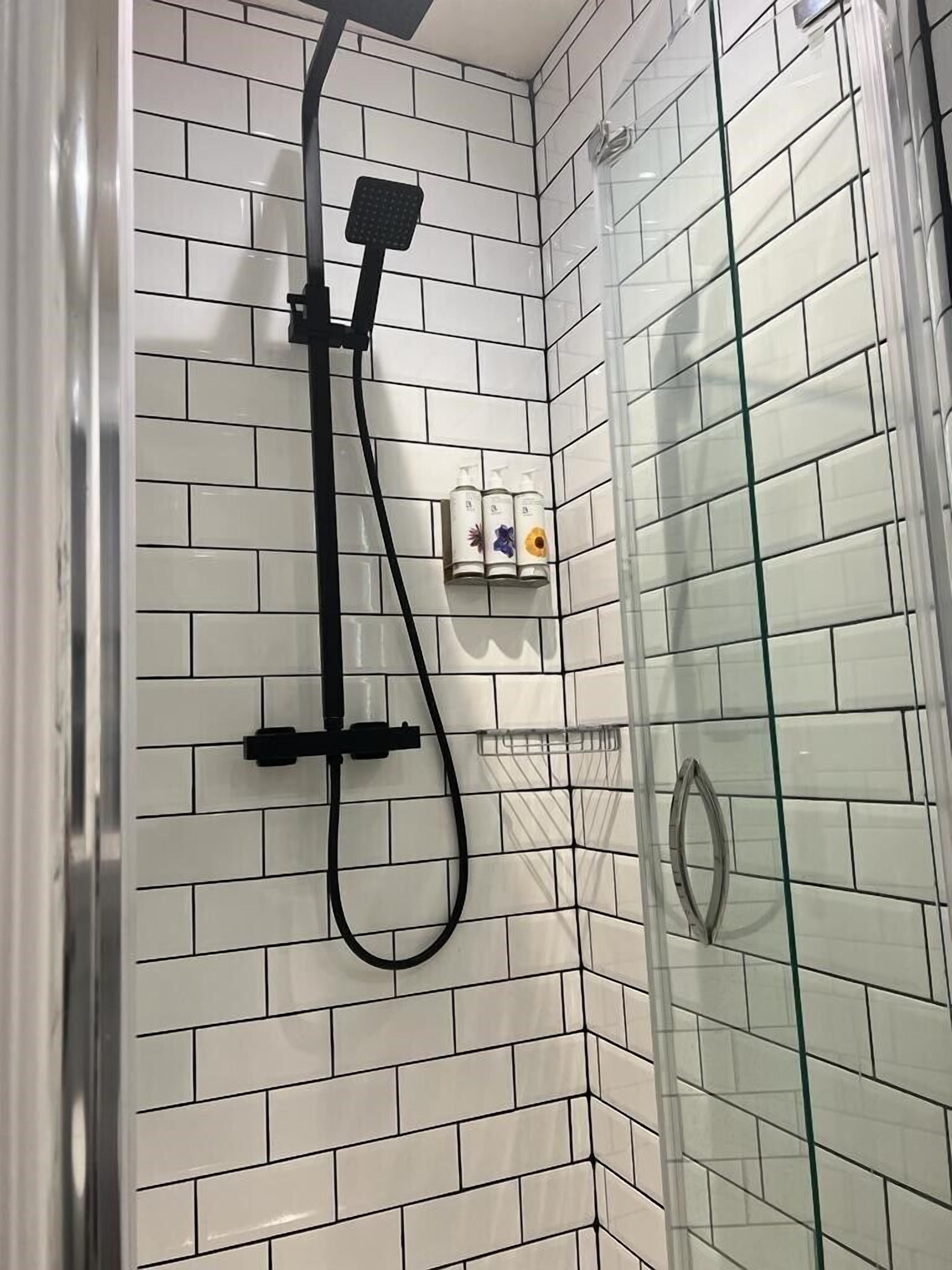A shower with a glass door, white tiled walls, and a black handheld showerhead.