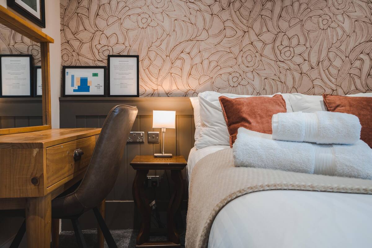 A hotel room with a bed, a desk, a chair, a nightstand with a lamp, and a wall with framed certificates.